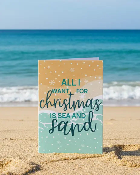 A Christmas greeting card with 'All I Want for Christmas is Sea and Sand' text, featuring a colorful graphic design with snowflakes and a sandy background, standing on a beach. 