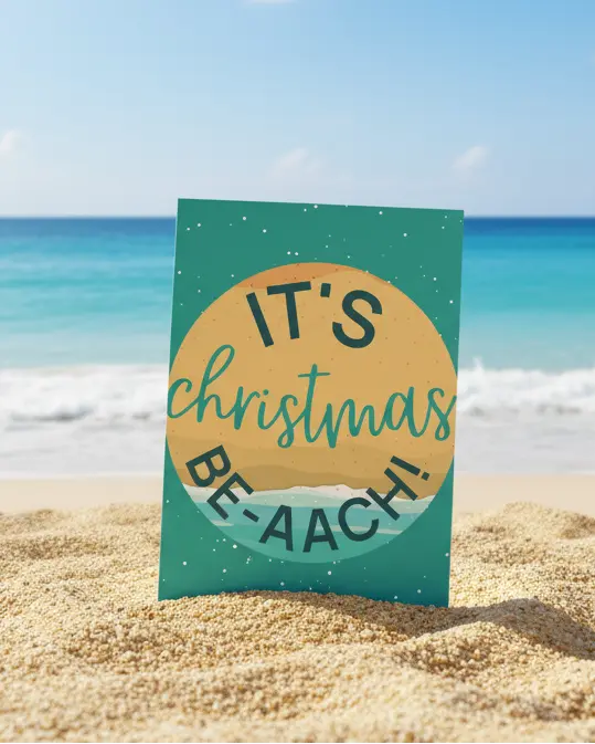 A Christmas greeting card with 'It's Christmas BEACH-AACH' printed on it, accompanied by a white kraft paper envelope, placed on a wooden surface with decorative elements in the background.