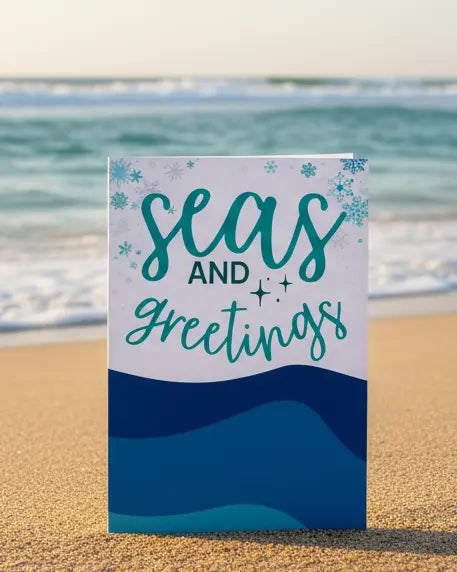 Seas And Greetings Christmas Card with snowflake and sea design sitting on a beach. 